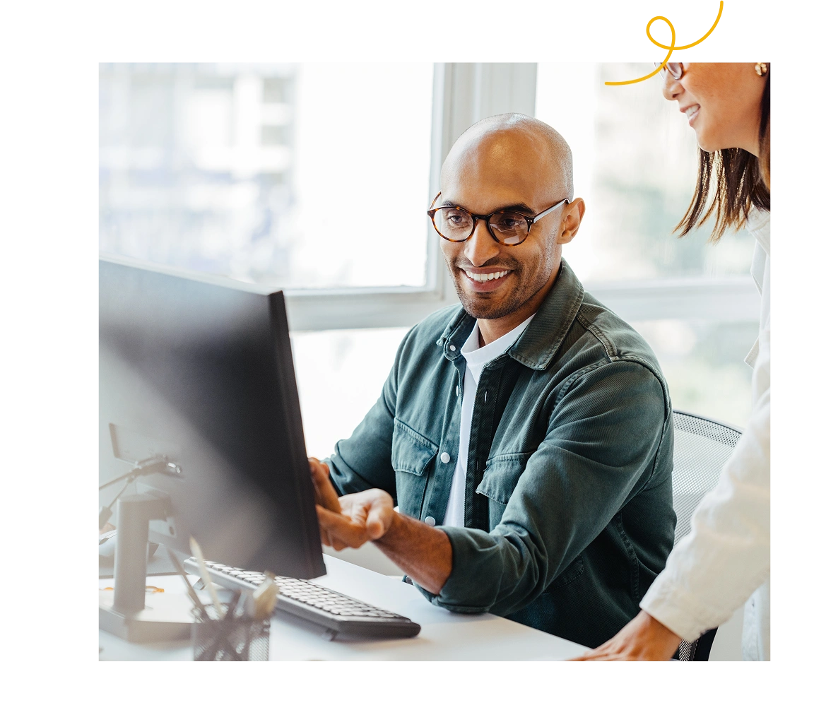 A smiling man wearing glasses works at a computer while discussing something with a colleague beside him