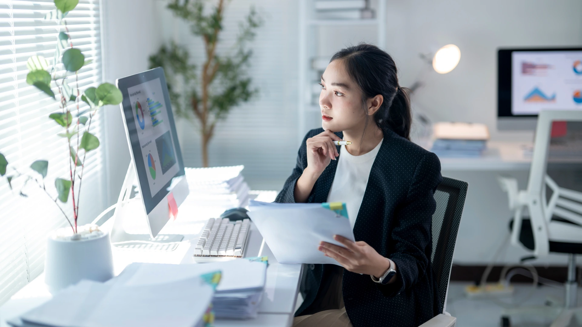 A woman is sitting at a desk with a computer monitor and a stack of papers in front of her.