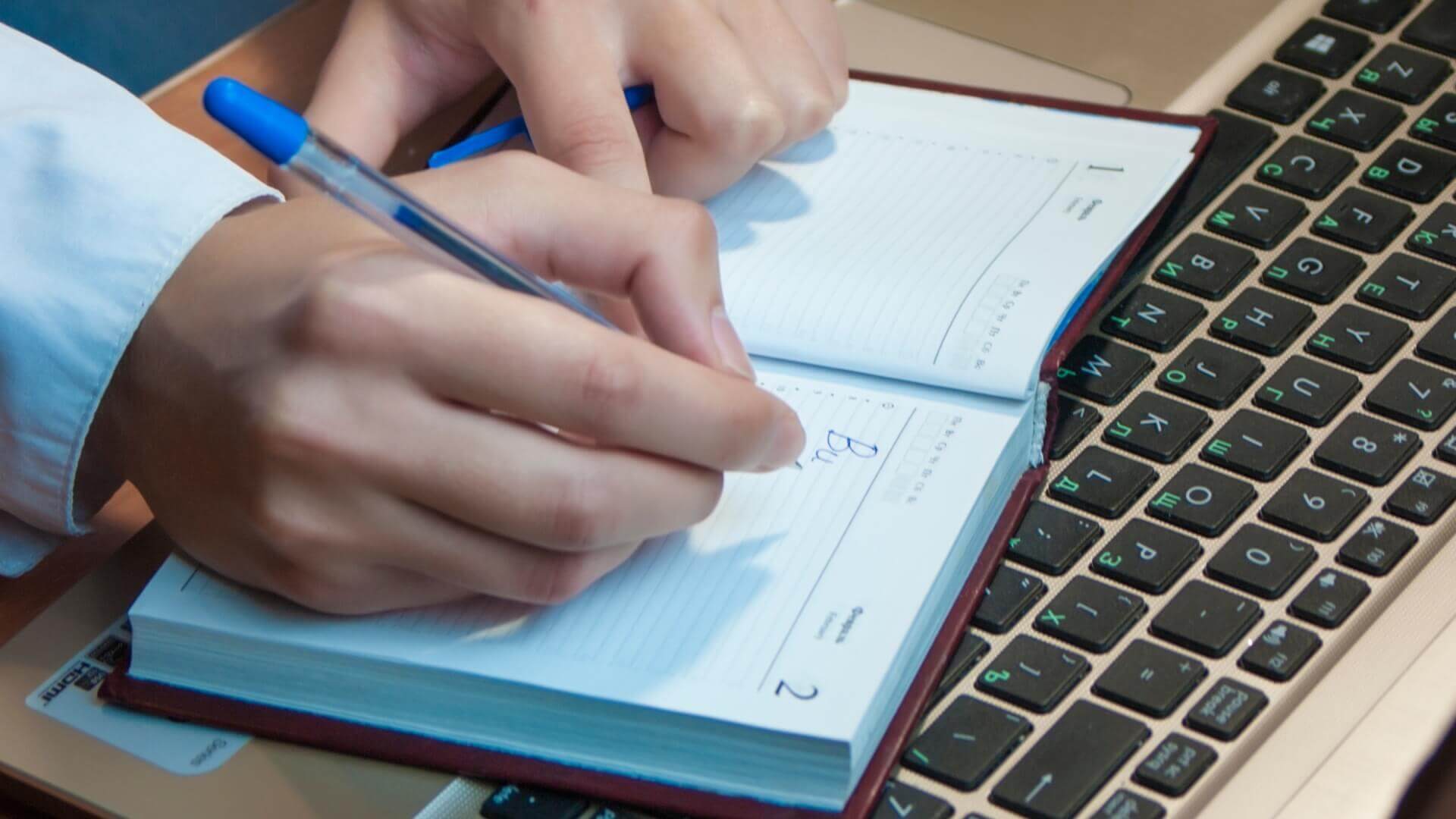 Person writing something in a notebook above the laptop