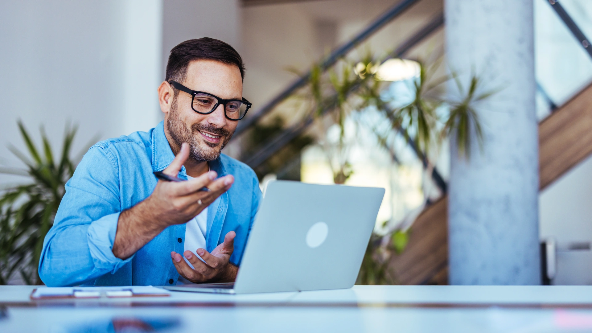 Smiling man using a laptop during an online conference call, sitting in a bright office space with plants and natural light, portraying communication, modern technology, and professional interaction