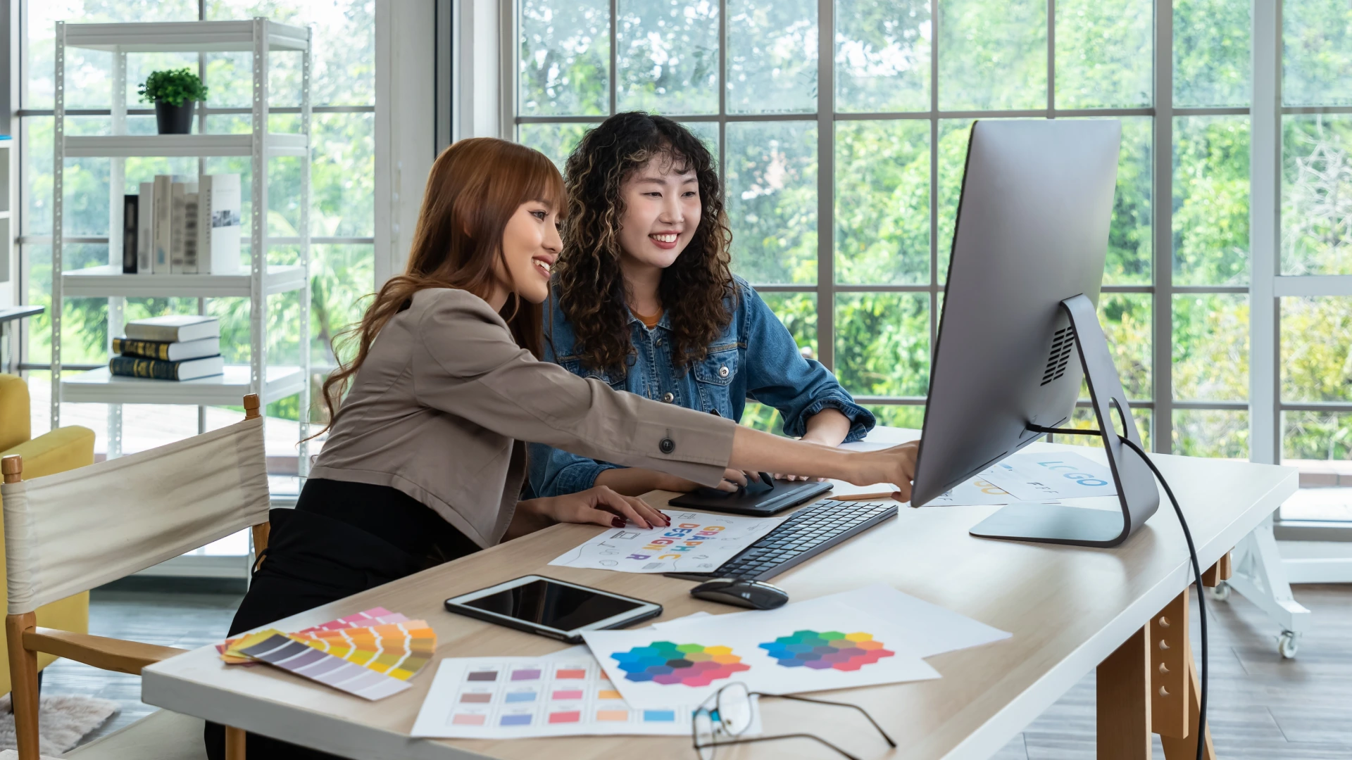 Two smiling young Asian female woman businessperson graphic designer working together, meeting and brainstorming, using computer in office, pointing at document.