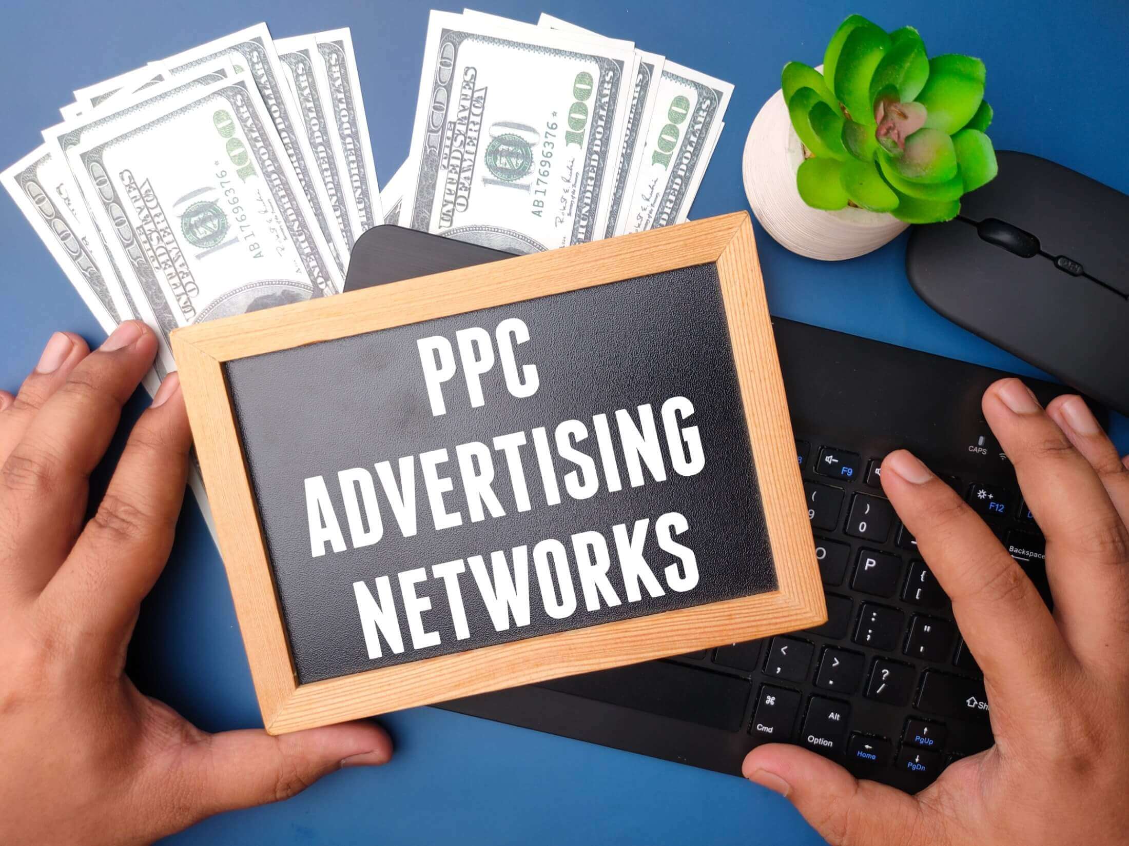 An image of a black-framed board with the words "PPC ADVERTISING NETWORKS" written on it, placed next to a wireless keyboard and a stack of banknotes, indicating a financial theme related to pay-per-click advertising.