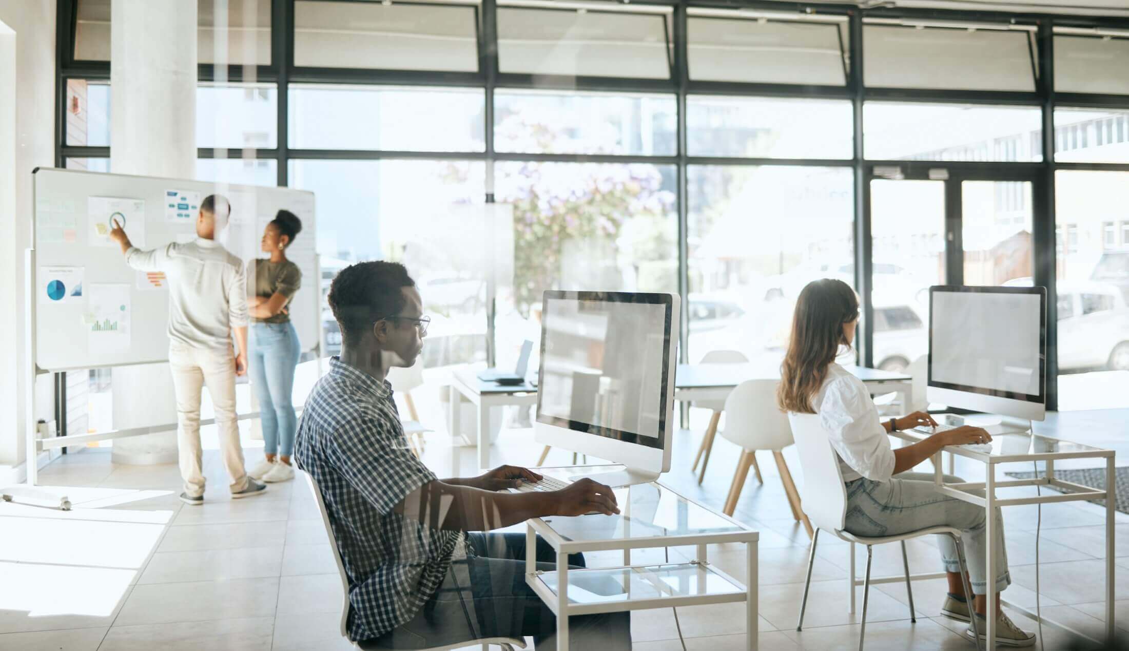 An image showing a modern office environment where diverse business professionals are working at their computers, some are engaged in a presentation on a whiteboard, depicting a collaborative workspace.