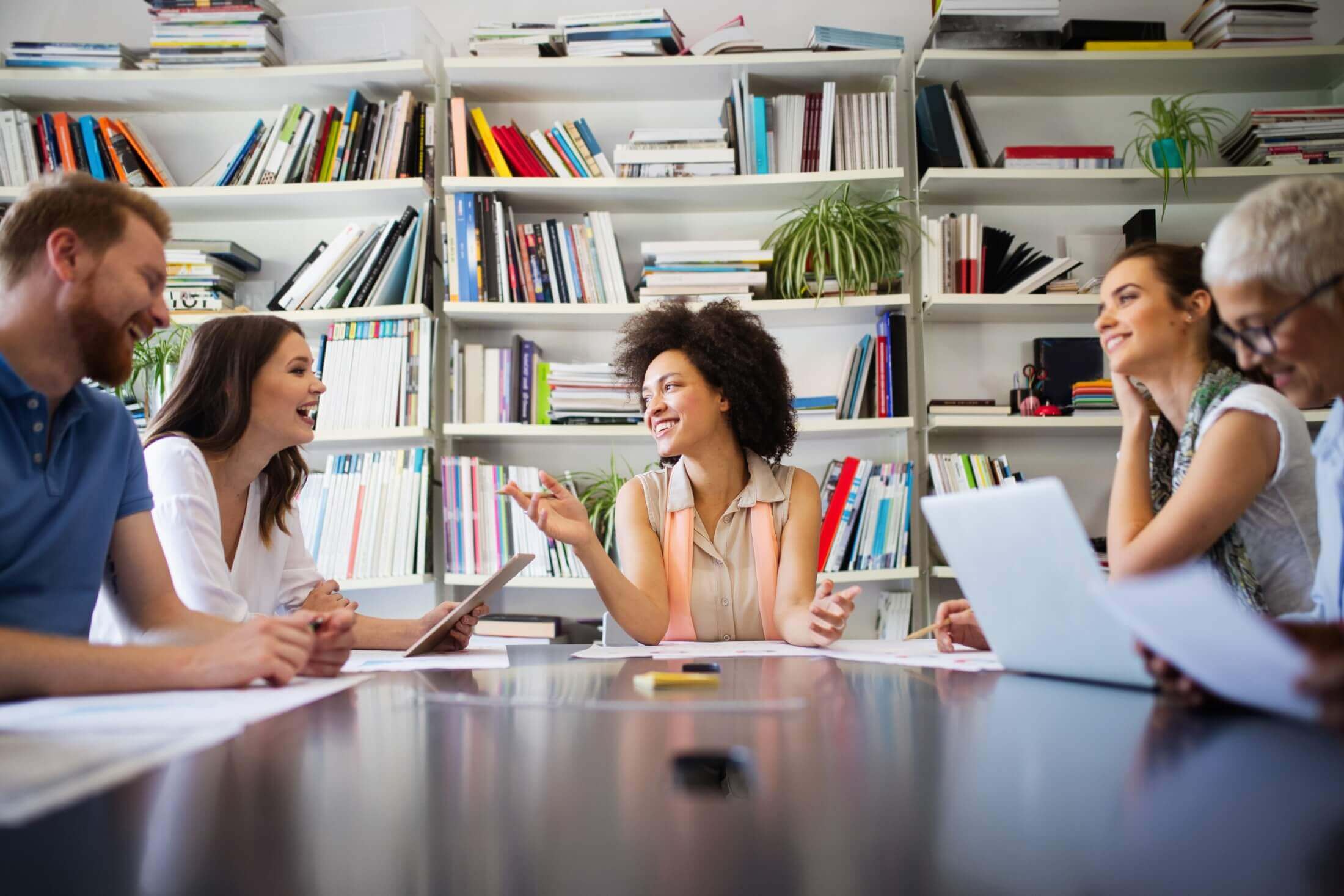 A group of business colleagues in a casual meeting within a library, with a woman gesturing during a creative brainstorming session.