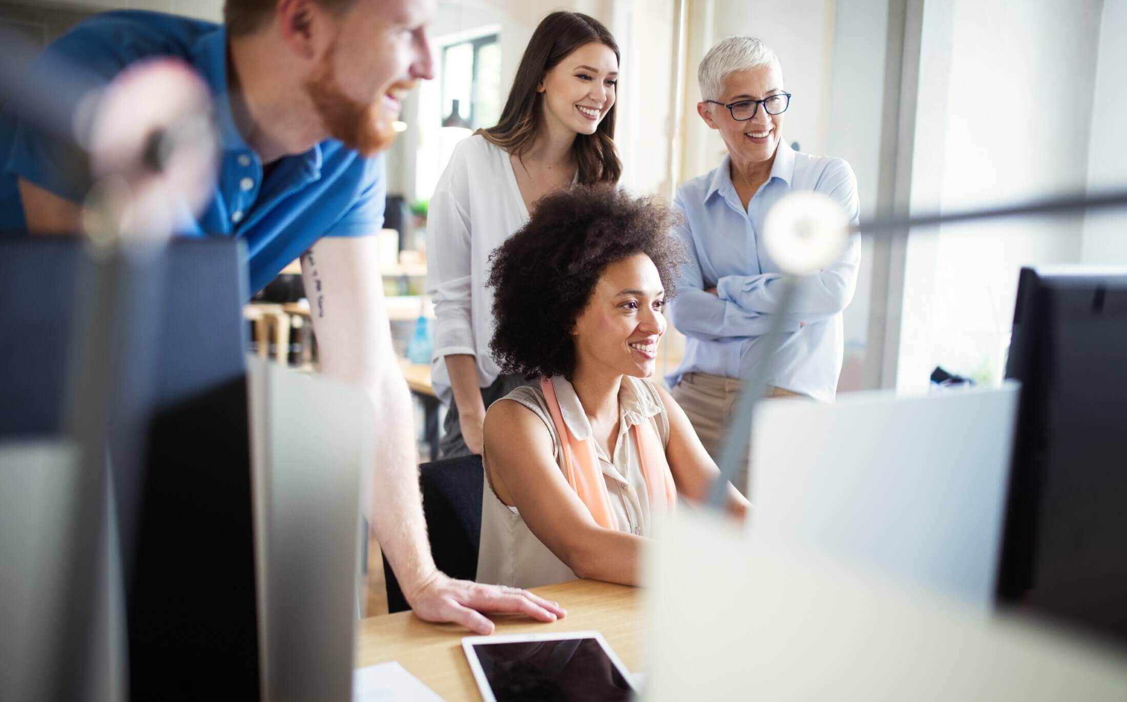 A diverse team of cheerful business professionals gathered around a computer in a bright office setting, engaging in a collaborative discussion.