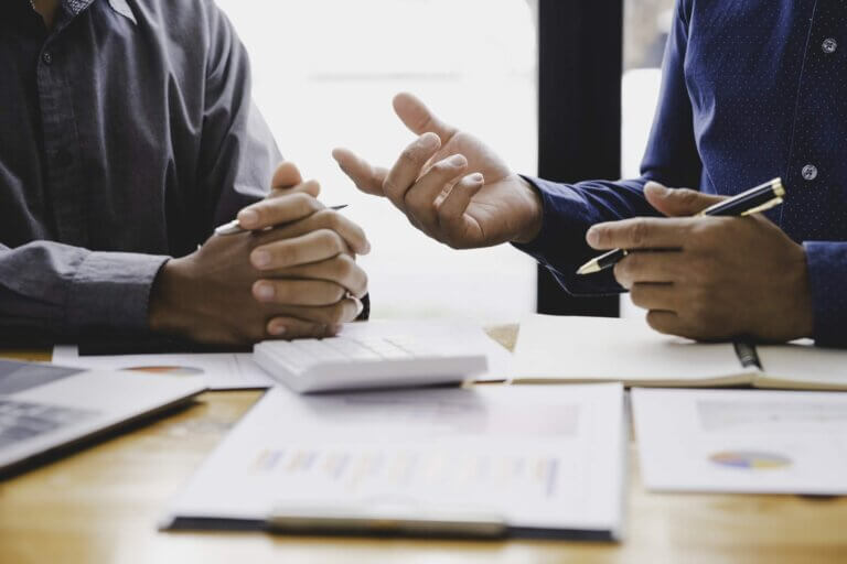 Two professionals in a business meeting, with focus on hands, discussing over documents with charts and a calculator on the table, symbolizing analysis of corporate risks and processes.