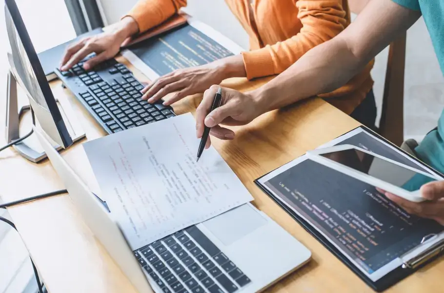 Two people collaborating on a coding project at a desk with laptops and documents