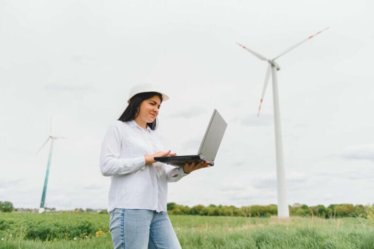 Female engineer in a white hard hat and casual attire using a laptop while standing in a field with wind turbines in the background, representing renewable energy and technology integration.