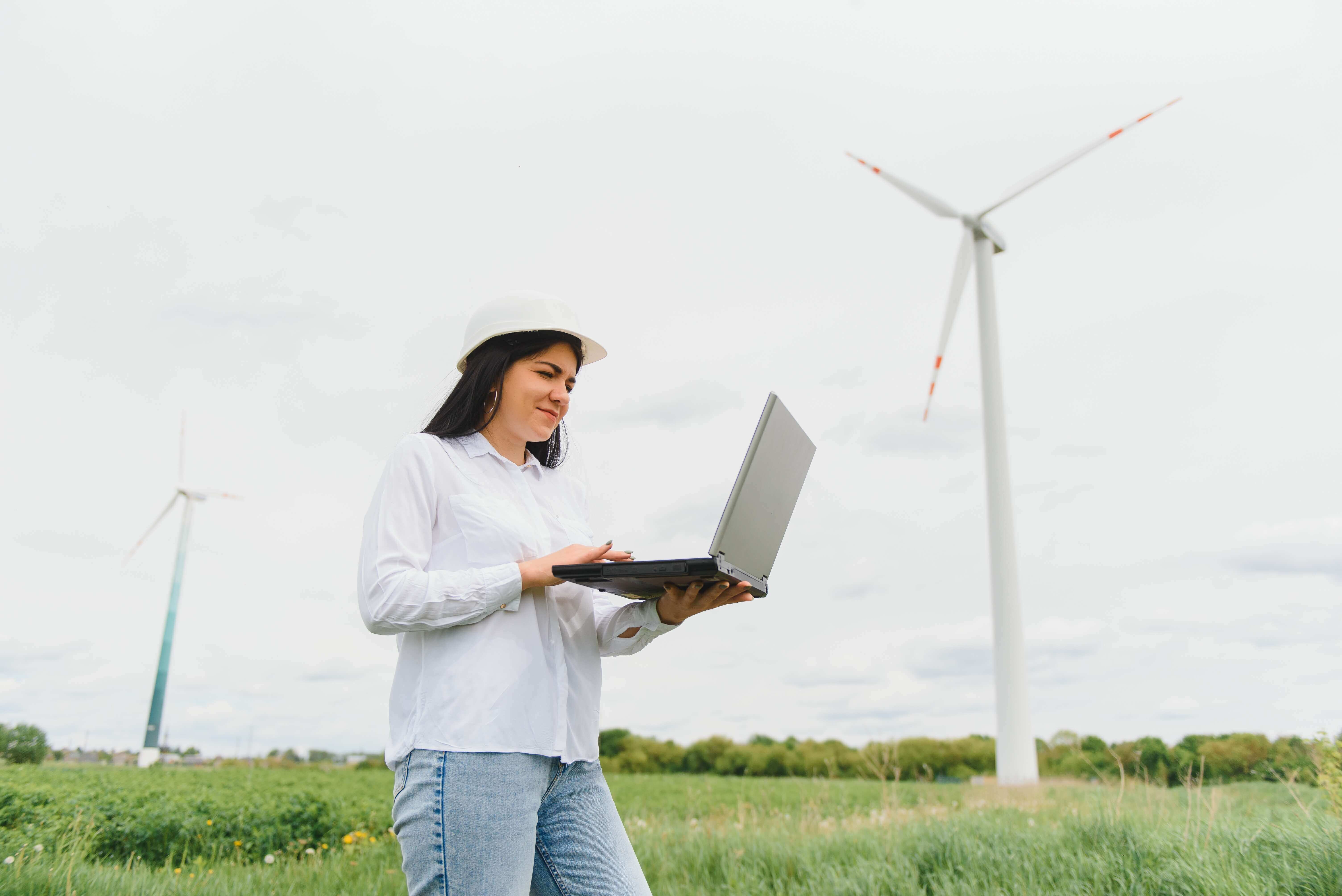 Female engineer in a white hard hat and casual attire using a laptop while standing in a field with wind turbines in the background, representing renewable energy and technology integration.