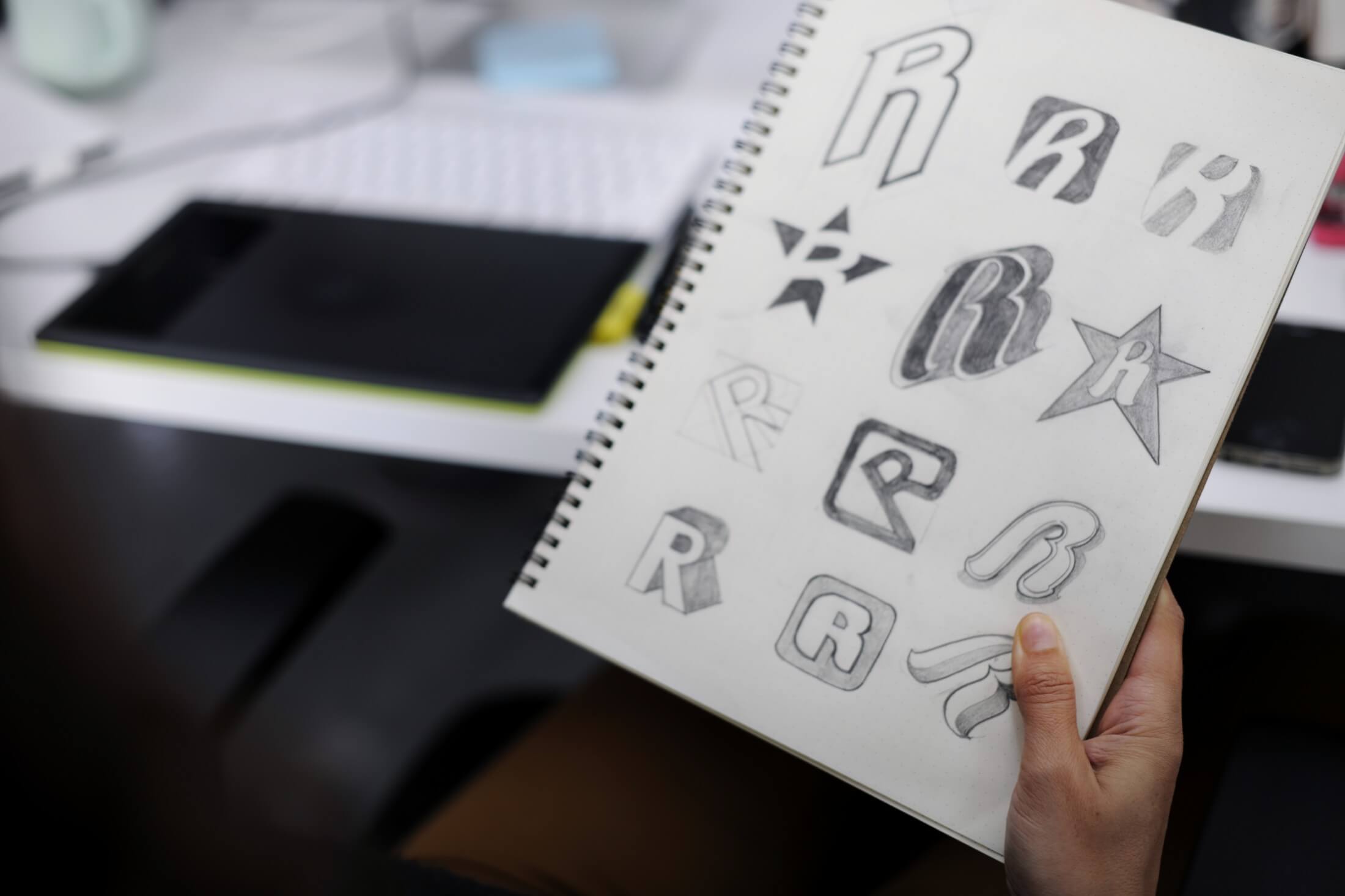 A hand holding open a sketchbook filled with various hand-drawn logo design concepts featuring the letter 'R'.
