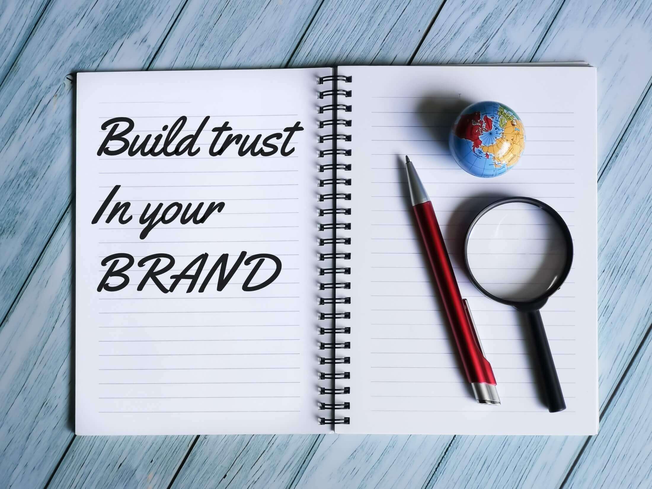 Notebook with inspirational branding message 'Build trust in your brand' alongside a magnifying glass and a miniature globe, emphasizing global brand integrity.