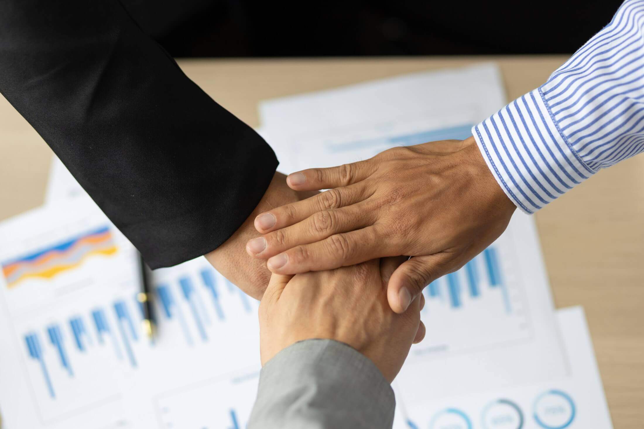 Two business individuals performing a handshake over a desk with financial documents, signifying a corporate partnership agreement.