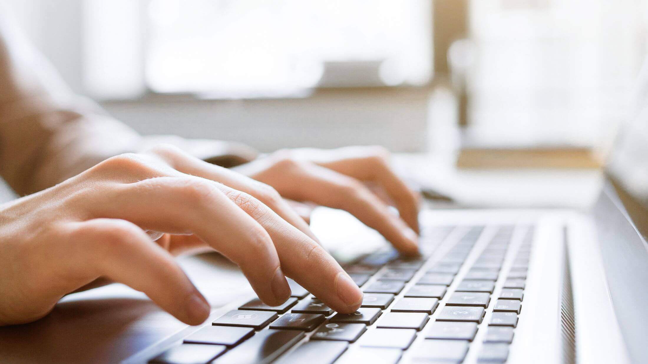Close-up of a person's hands typing on a laptop keyboard, with a blurred background suggesting a bright and airy room.