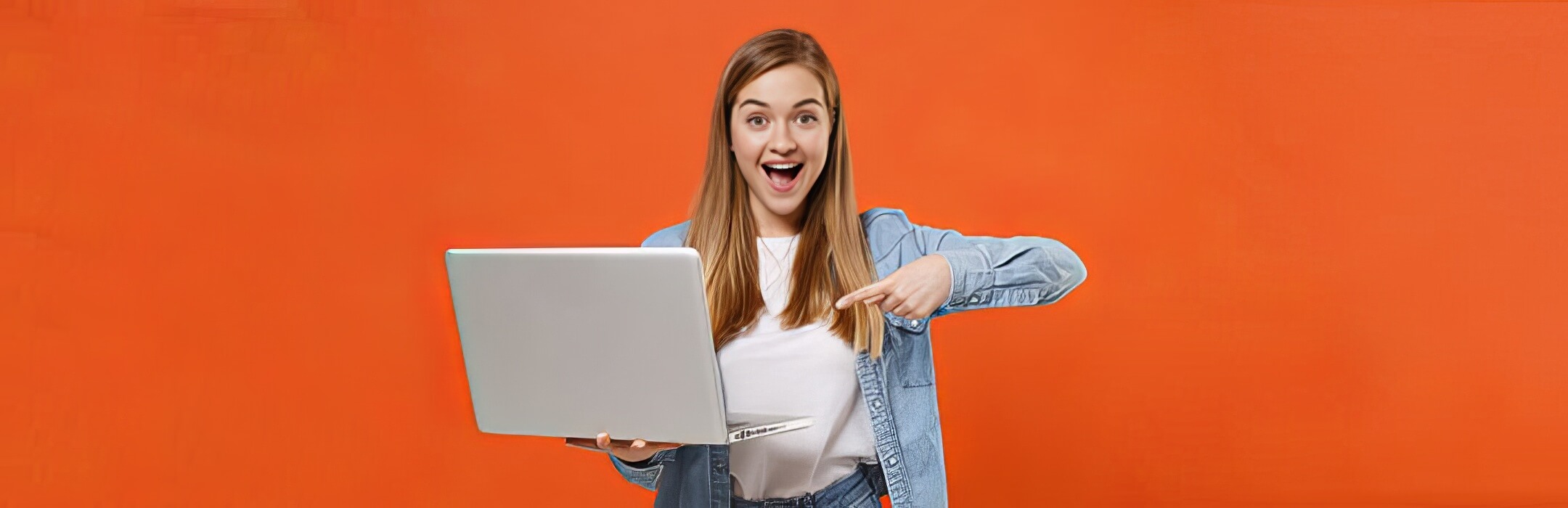 Excited young woman girl in casual denim clothes posing isolated on orange wall background studio portrait. People lifestyle concept. Mock up copy space. Pointing index finger on laptop pc computer