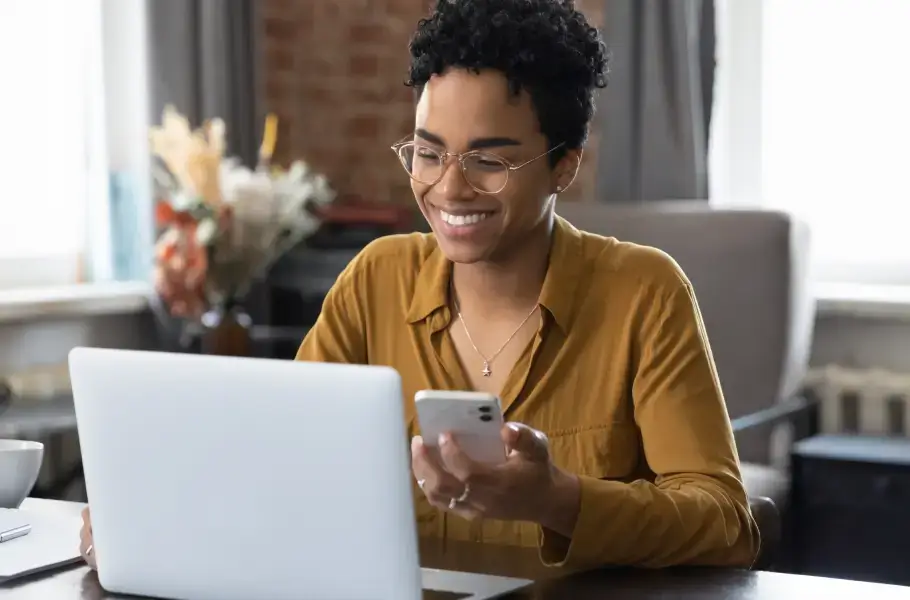 Woman smiling while using a laptop and holding a smartphone