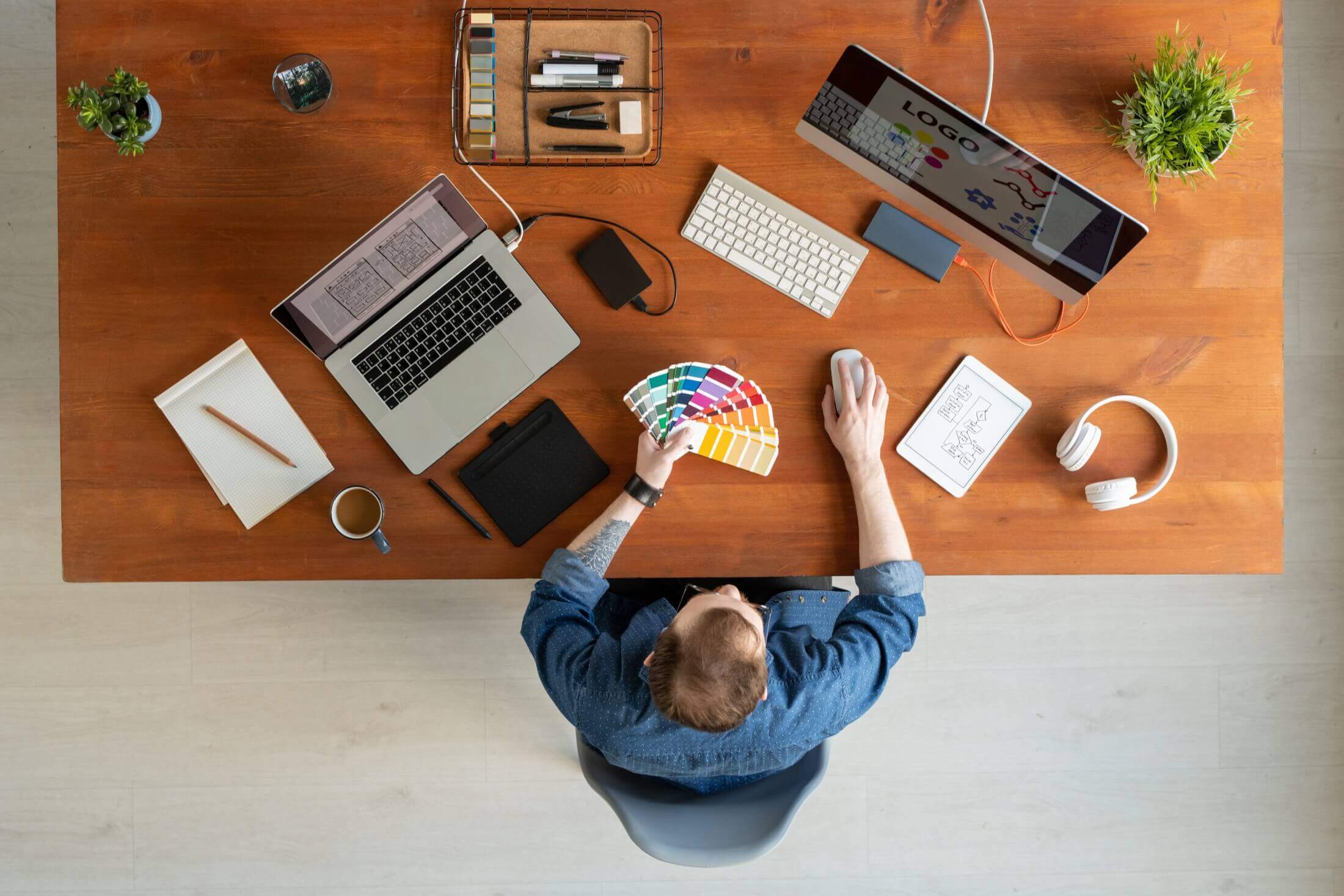 Overhead view of a designer at a wooden desk with various branding and identity design elements, such as color palettes and mockups, while holding a color sampler.