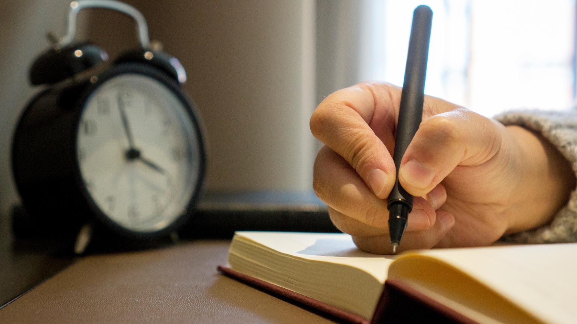 writing in a notebook with an alarm clock beside it